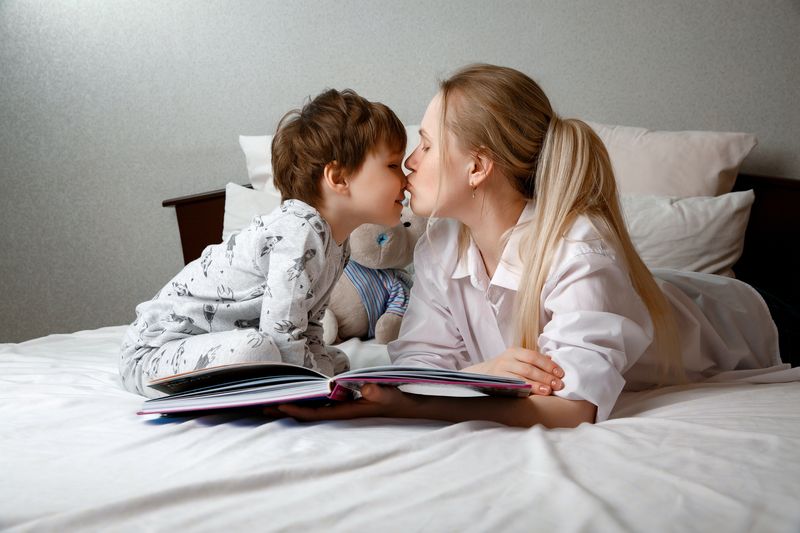 Süßer Vorschulkindersohn hält ein Buch, liest Märchen für Mama, liegt zusammen im Bett, glückliche Mutter ...