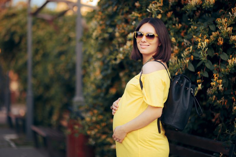 Glückliche schwangere Frau mit Sonnenbrille und Rucksack. Mutter redet einen Spaziergang im Park ...