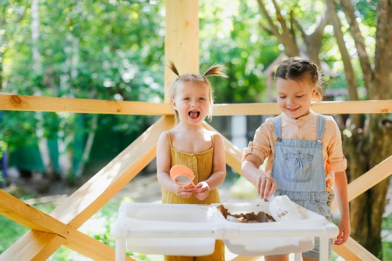 Glückliche Schwesterkinder spielen mit Sand und Wasser in sensorischen Körben auf dem sensorischen Tisch im Freien, sensorischen ...