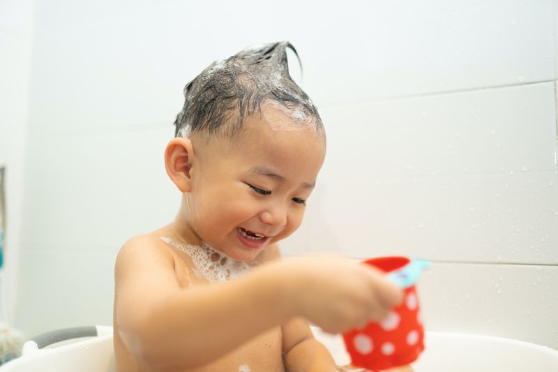 lächelnder asiatischer Babyjunge spielt mit Spielzeug an der Badewanne im Badezimmer, süßer asiatischer Kleinkindjunge mit lustigem...