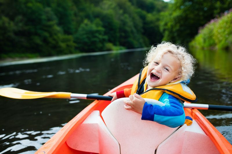 Glückliches Kind, das eine Kajakfahrt auf einem wunderschönen Fluss genießt. Kleiner gelockter Kleinkindjunge, der an heißem Sommertag Kajak fährt ...