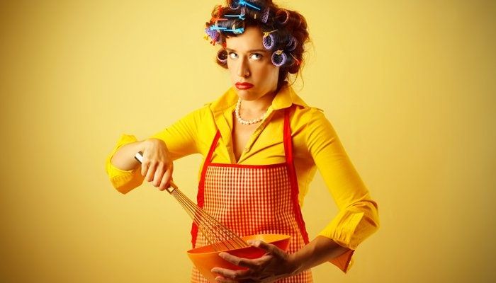 Eine Frau posiert im Hausfrauen-Outfit mit einem genervten Gesichtsausdruck.
